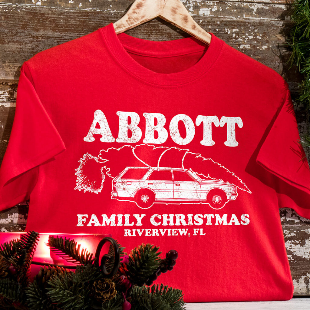 A red An Adventurous Christmas tee on a wooden backdrop surrounded by Christmas foliage