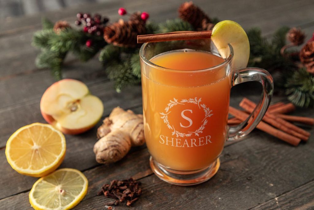 Wassail - a timeless drink. Tea and fruit based mocktail in a glass coffee mug, garnished with lemon, ginger, cinnamon, and apple.