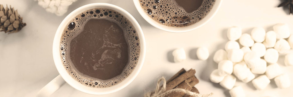 A cup of hot coffee surrounded by marshmallows, pine cones, and bundled sticks.